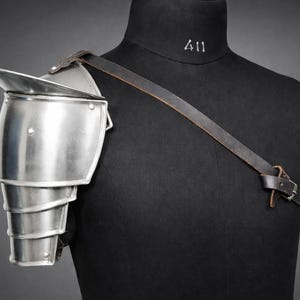 May include: A silver-colored shoulder pauldron, part of a suit of armor, is displayed on a black mannequin. The pauldron has a layered design and is secured with a brown leather strap. The mannequin has the number "411" printed on the neck.
