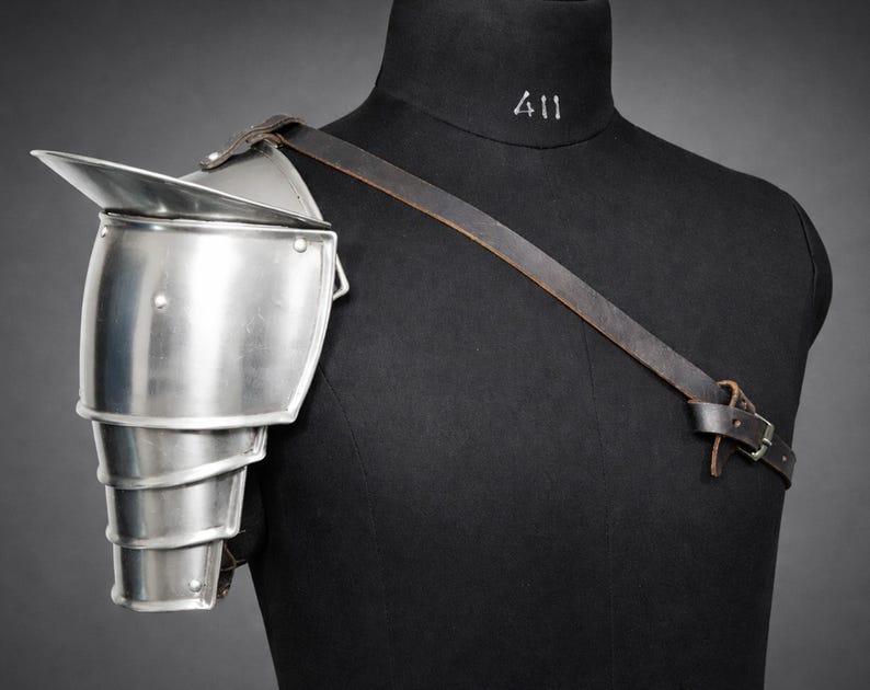 May include: A silver-colored shoulder pauldron, part of a suit of armor, is displayed on a black mannequin. The pauldron has a layered design and is secured with a brown leather strap. The mannequin has the number "411" printed on the neck.