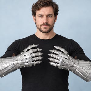 May include: A pair of silver-toned metal gauntlets featuring intricate embossed scrollwork patterns, articulated finger plates, and pointed claw-like tips extending from each finger, covering the hands and forearms in a protective armour style.