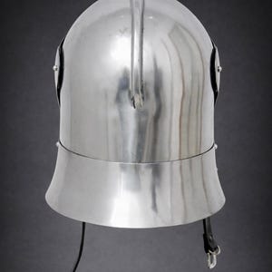 May include: A polished silver-colored metal helmet featuring a rounded dome, a central vertical ridge, and a flared lower section. The helmet includes two side rivets and a black leather chin strap with a metal buckle attached at the base.