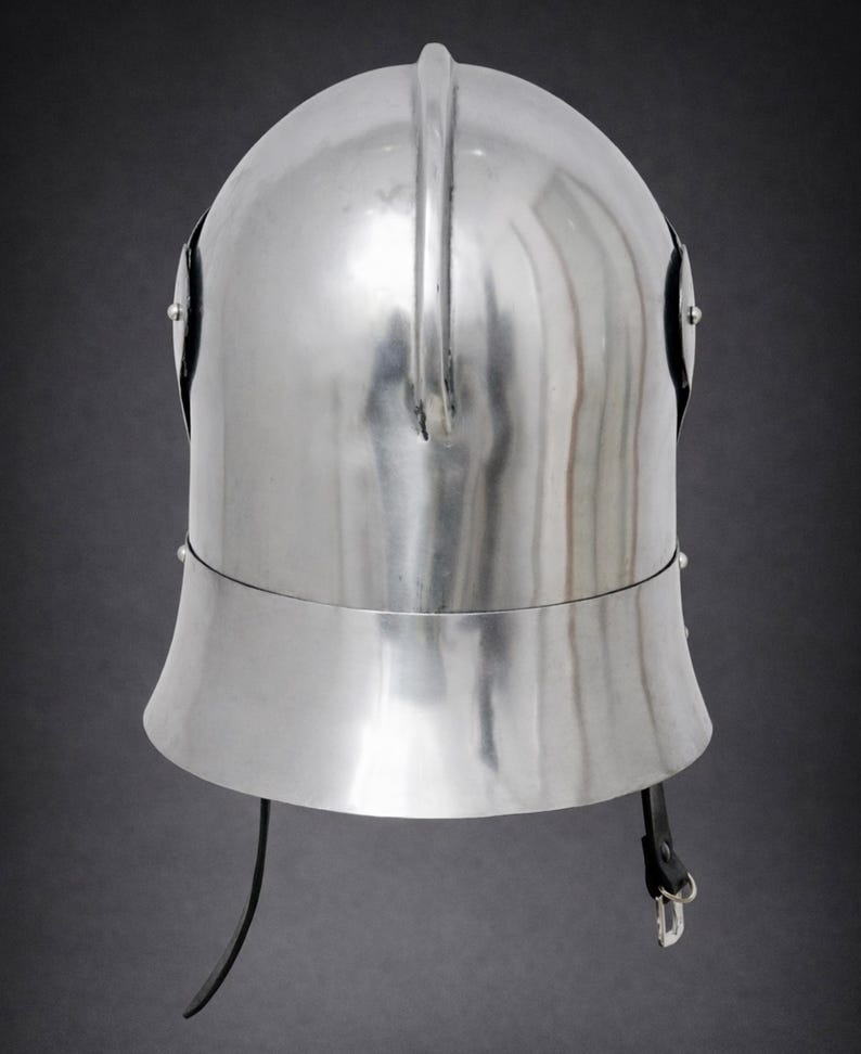 May include: A polished silver-colored metal helmet featuring a rounded dome, a central vertical ridge, and a flared lower section. The helmet includes two side rivets and a black leather chin strap with a metal buckle attached at the base.