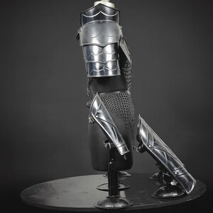 Puede incluir: Un conjunto de armadura de metal plateado pulido que incluye una hombrera segmentada, una t&uacute;nica de cota de malla y dos grebas a juego. Las piezas de la armadura se exhiben sobre un torso de maniqu&iacute; negro y soportes separados sobre fondo oscuro.
