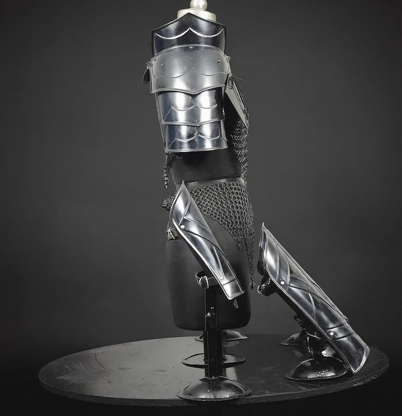 Puede incluir: Un conjunto de armadura de metal plateado pulido que incluye una hombrera segmentada, una t&uacute;nica de cota de malla y dos grebas a juego. Las piezas de la armadura se exhiben sobre un torso de maniqu&iacute; negro y soportes separados sobre fondo oscuro.