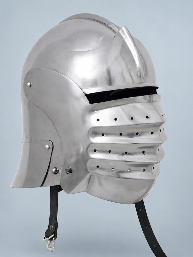 May include: A polished silver-colored metal helmet featuring a rounded dome, a prominent central ridge, and a hinged visor with horizontal ridges and multiple ventilation holes. The helmet includes black leather chin straps with metal buckles.