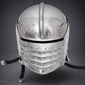 May include: A polished silver-colored metal knight helmet featuring a rounded dome, a horizontal eye slit, and a lower visor section with multiple ventilation holes. Black leather straps with silver-toned metal buckles are attached to the sides.
