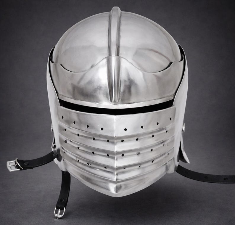May include: A polished silver-colored metal knight helmet featuring a rounded dome, a horizontal eye slit, and a lower visor section with multiple ventilation holes. Black leather straps with silver-toned metal buckles are attached to the sides.