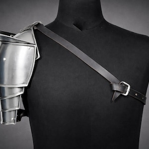 May include: A silver-colored shoulder pauldron made of metal, with layered segments and a pointed top. It is attached to a black leather strap that buckles across the chest, displayed on a black mannequin.