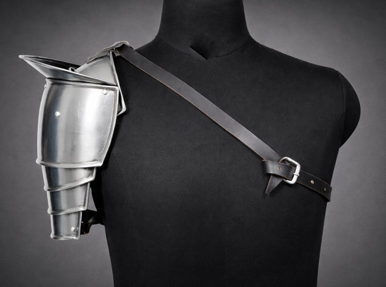 May include: A silver-colored shoulder pauldron made of metal, with layered segments and a pointed top. It is attached to a black leather strap that buckles across the chest, displayed on a black mannequin.