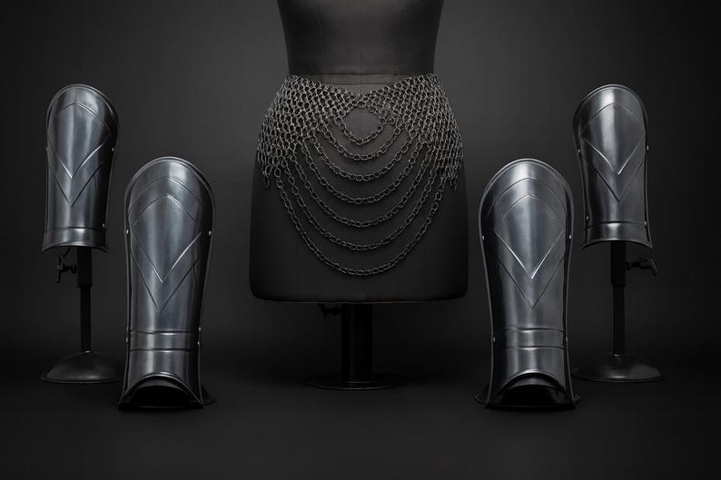 Puede incluir: Un conjunto de piezas de armadura de metal oscuro que incluye cuatro grebas con detalles en forma de chevr&oacute;n y un cintur&oacute;n de cota de malla con m&uacute;ltiples capas drapeadas de anillos de metal entrelazados sobre fondo oscuro.