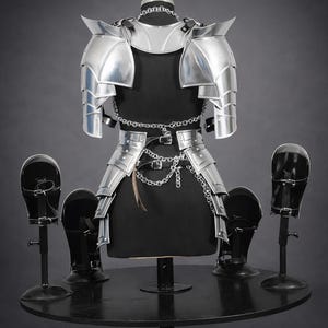 K&ouml;nnte beinhalten: Ein Set aus polierter, silberfarbener Metallr&uuml;stung, bestehend aus einem segmentierten Schulter- und Torsoharnisch mit integrierten silbernen Ketten und Schnallen, pr&auml;sentiert neben vier passenden, geschwungenen Bein- oder Armschienen auf schwarzen St&auml;ndern.