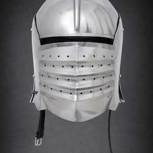 May include: A polished silver-colored metal knight helmet featuring a rounded top, a horizontal eye slit, and a lower visor section with three rows of small ventilation holes. A black leather chin strap with a metal buckle hangs from the side.