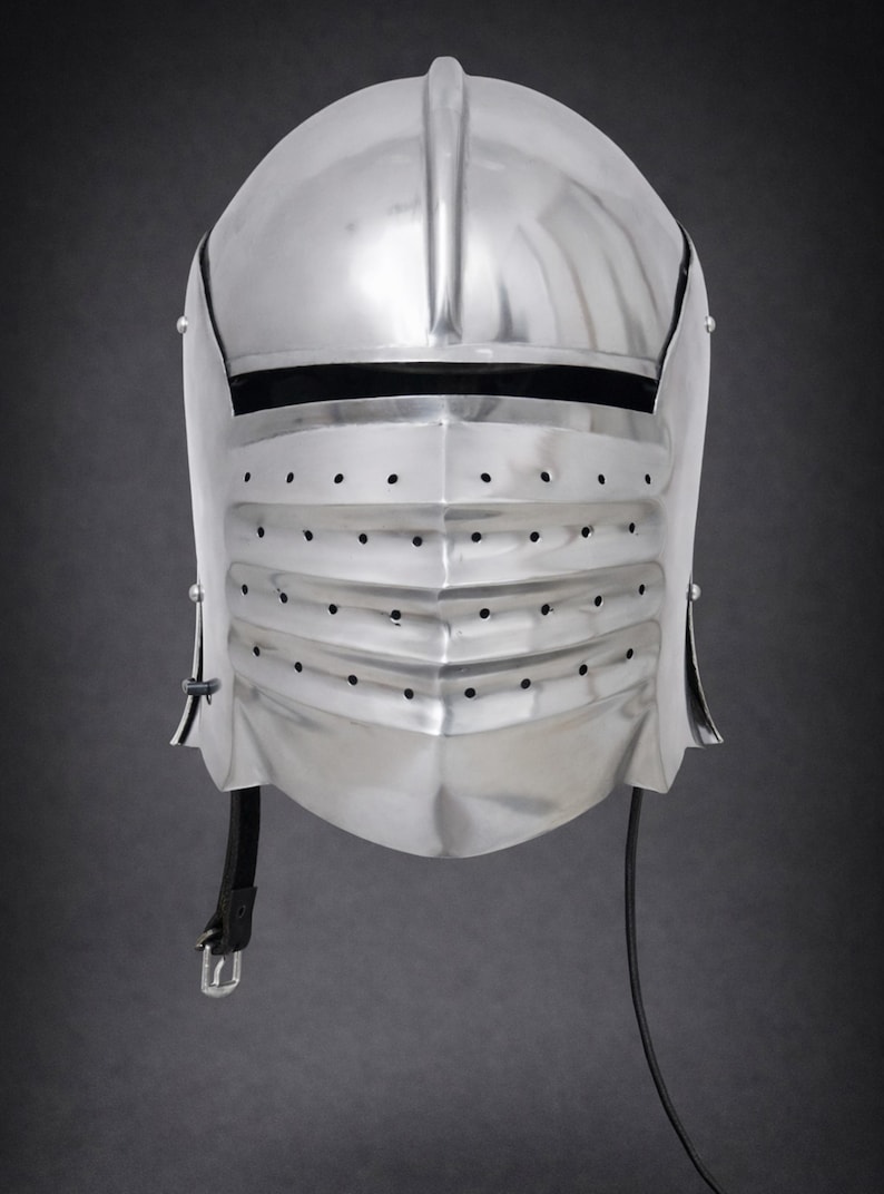 May include: A polished silver-colored metal knight helmet featuring a rounded top, a horizontal eye slit, and a lower visor section with three rows of small ventilation holes. A black leather chin strap with a metal buckle hangs from the side.