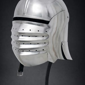 May include: A polished silver-colored metal helmet featuring a rounded dome, a horizontal eye slit, and a protruding visor with three horizontal ridges containing small ventilation holes. A black leather chin strap with a metal buckle hangs below.