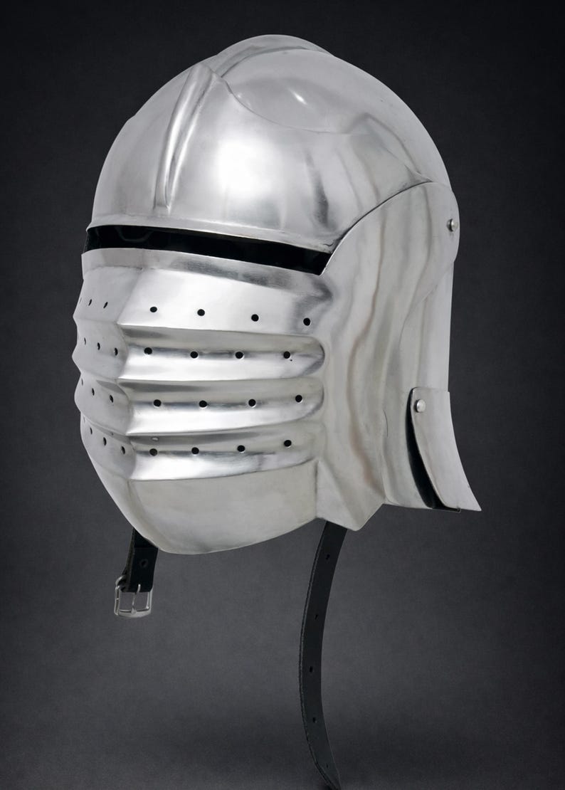 May include: A polished silver-colored metal helmet featuring a rounded dome, a horizontal eye slit, and a protruding visor with three horizontal ridges containing small ventilation holes. A black leather chin strap with a metal buckle hangs below.