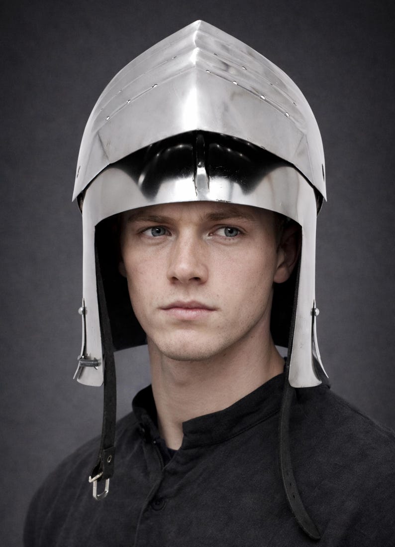 May include: A polished silver-colored metal bascinet helmet featuring a pointed visor and a rounded skull cap. The helmet includes a hinged visor mechanism with visible rivets along the edges and black leather chin straps hanging down on both sides.