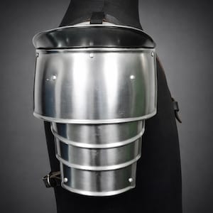 May include: A polished steel shoulder pauldron, part of medieval armor, with a black leather strap and trim. The pauldron is composed of multiple articulated plates for flexibility and protection. Rivets secure the plates.