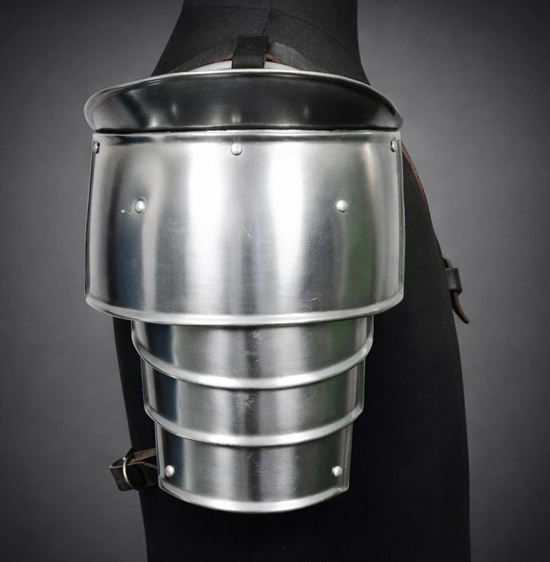 May include: A polished steel shoulder pauldron, part of medieval armor, with a black leather strap and trim. The pauldron is composed of multiple articulated plates for flexibility and protection. Rivets secure the plates.
