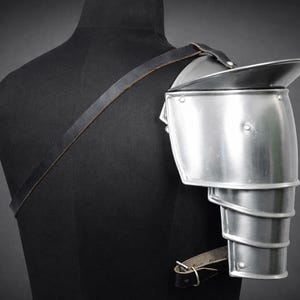 May include: A silver-colored metal shoulder pauldron, part of a suit of armor, is displayed on a black mannequin. The pauldron has a layered design for the arm and a large, curved plate to protect the shoulder. A leather strap secures it.