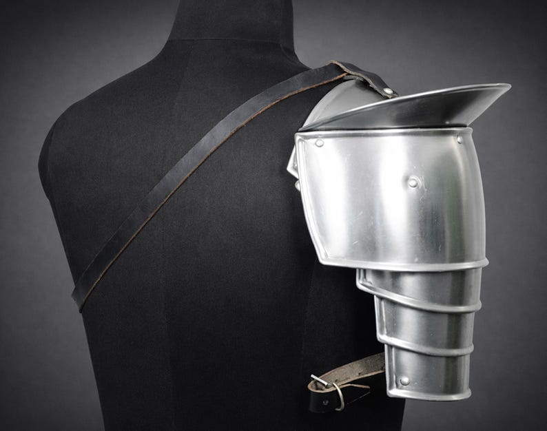 May include: A silver-colored metal shoulder pauldron, part of a suit of armor, is displayed on a black mannequin. The pauldron has a layered design for the arm and a large, curved plate to protect the shoulder. A leather strap secures it.