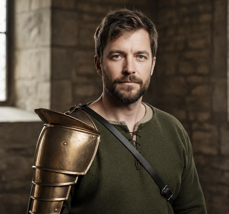 May include: A man wearing a bronze shoulder armor piece and a dark green tunic. The armor is made of overlapping plates and is attached to a black leather strap. The man has a beard and is looking at the camera.