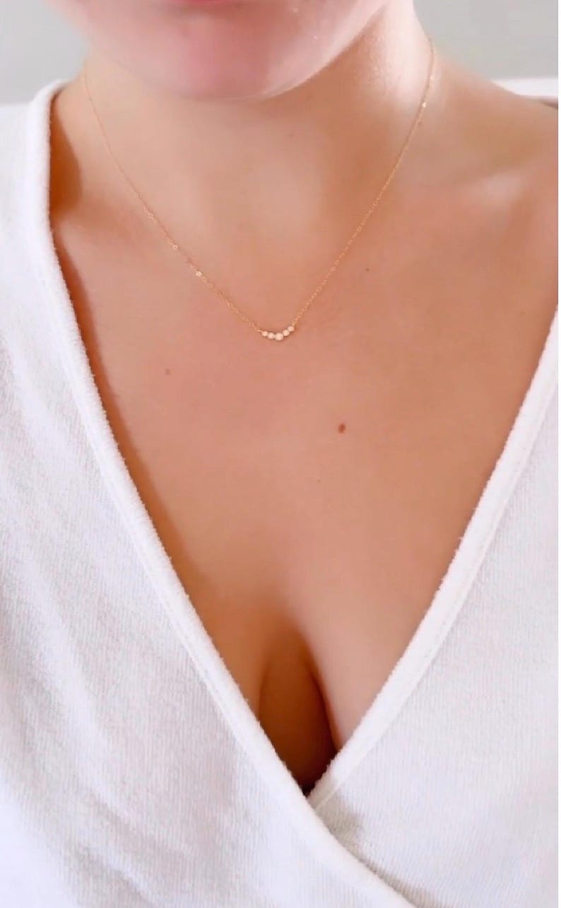 May include: A delicate gold chain necklace featuring a curved bar pendant set with five small, round, clear stones arranged in a graduated size pattern. The necklace rests against the collarbone, showcasing a thin, polished metal link chain.