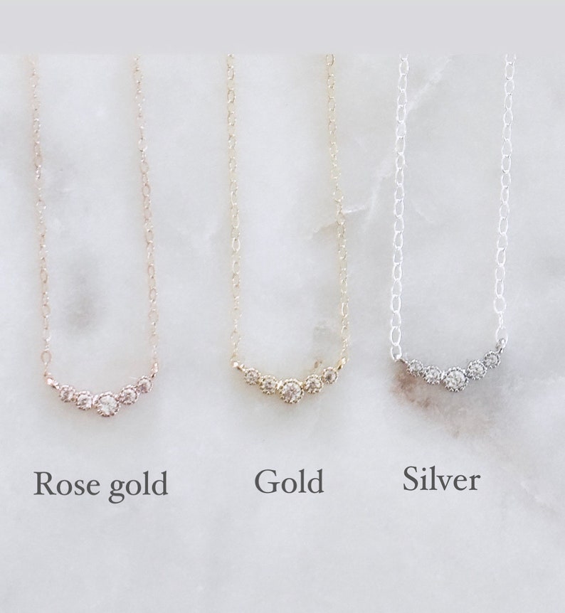 May include: Three delicate necklaces featuring a curved bar of five small round stones. The left necklace is rose gold, the center is gold, and the right is silver. Each necklace has a thin chain and is labeled below as Rose gold, Gold, and Silver.