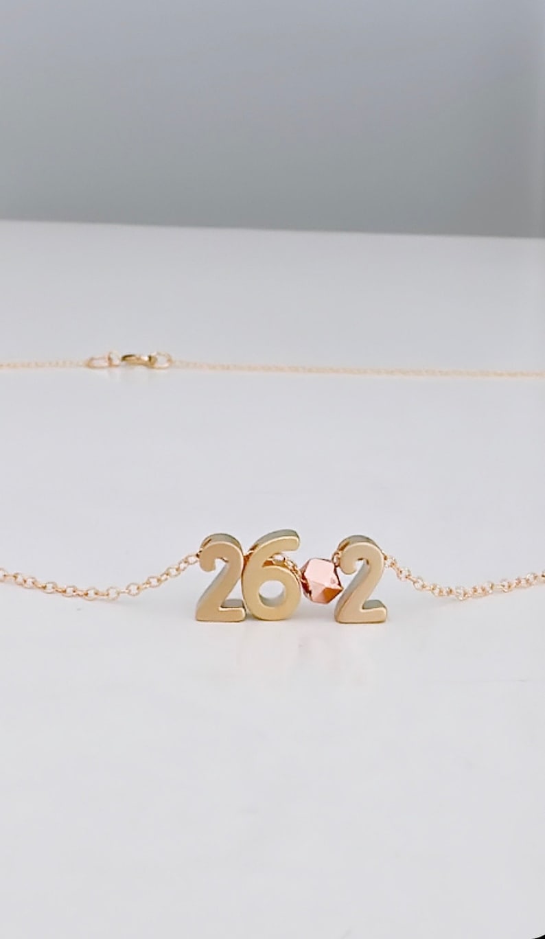May include: Gold necklace with a pendant featuring the numbers 262. A small rose gold bead separates the numbers.