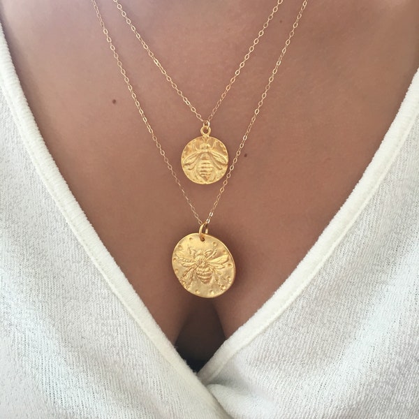 Könnte beinhalten: Eine goldene Kettenkette mit zwei bienenförmigen Anhängern. Der größere Anhänger befindet sich am unteren Ende der Kette und der kleinere Anhänger darüber.