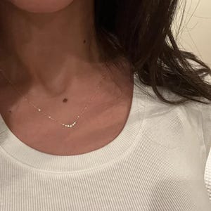 May include: A delicate gold chain necklace featuring a small, curved pendant set with a cluster of five tiny, clear, round-cut stones. The necklace rests against the neck, showcasing a minimalist design with a thin, polished metal link chain.