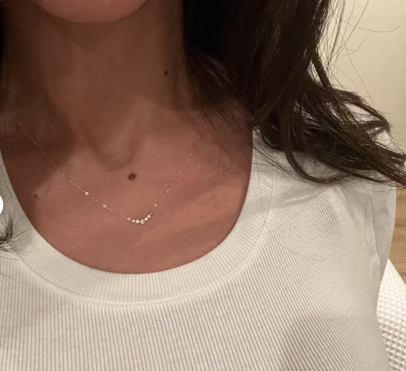 May include: A delicate gold chain necklace featuring a small, curved pendant set with a cluster of five tiny, clear, round-cut stones. The necklace rests against the neck, showcasing a minimalist design with a thin, polished metal link chain.