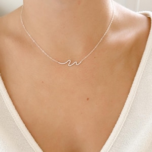 Puede incluir: Un delicado collar de plata con un colgante en forma de ola. El collar se lleva alrededor del cuello. El diseño de la ola es una pieza de joyería sencilla y elegante. Una joya delicada.