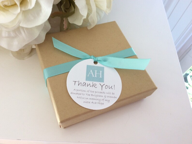 Puede incluir: Una caja de regalo marr&oacute;n atada con una cinta azul claro y una etiqueta circular blanca con las iniciales "AH" en un cuadrado azul claro. La etiqueta dice "&iexcl;Gracias! Una parte de las ganancias se donar&aacute; a The Brigham & Women NICH en memoria de mi sobrina Ava Hope."
