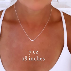 May include: A silver-toned necklace featuring a delicate chain and a central pendant arrangement of seven small cubic zirconia stones. The necklace has a total length of 18 inches and includes 7 cz stones as indicated by the text in the image.