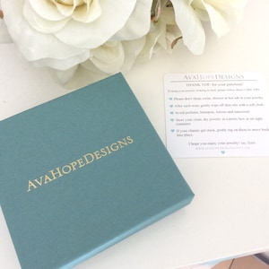 May include: A square teal jewelry box with the gold text AVAHOPEDESIGNS printed on the lid. Next to the box is a white rectangular card with the text AVAHOPEDESIGNS and care instructions for jewelry maintenance printed in black and blue ink.