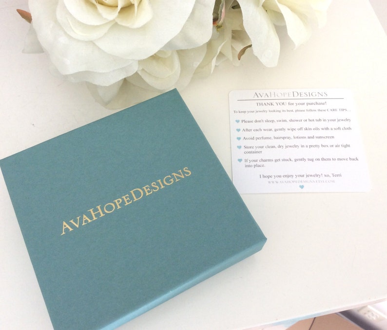 May include: A square teal jewelry box with the gold text AVAHOPEDESIGNS printed on the lid. Next to the box is a white rectangular card with the text AVAHOPEDESIGNS and care instructions for jewelry maintenance printed in black and blue ink.