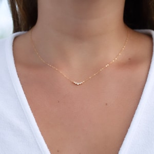 May include: A delicate gold chain necklace featuring a small, V-shaped pendant set with five tiny, clear, round-cut stones arranged in a graduated arc. The thin, polished gold chain rests against the collarbone, creating a subtle, minimalist look.