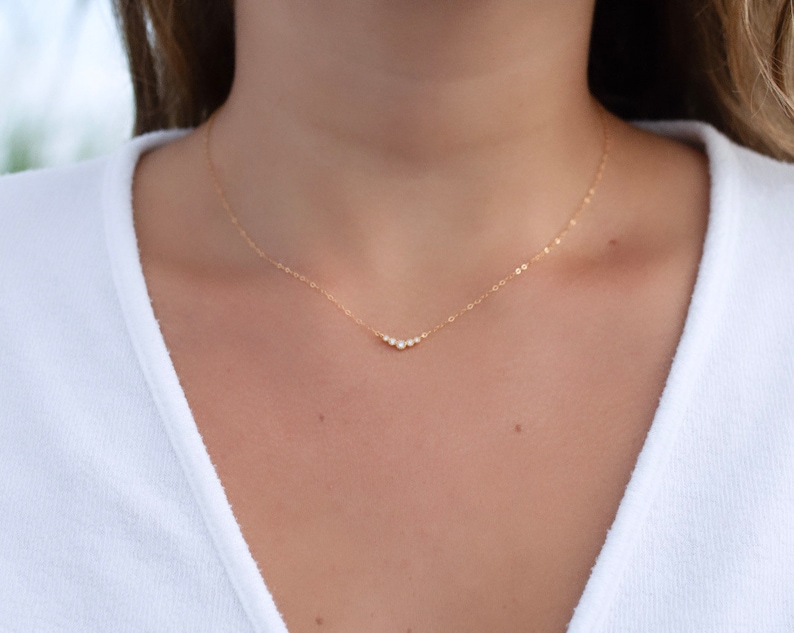 May include: A delicate gold chain necklace featuring a small, V-shaped pendant set with five tiny, clear, round-cut stones arranged in a graduated arc. The thin, polished gold chain rests against the collarbone, creating a subtle, minimalist look.