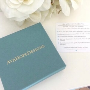 May include: A teal blue box with the text "AvaHopeDesigns" in gold lettering. A white card with the text "AvaHopeDesigns" and care instructions for jewelry.