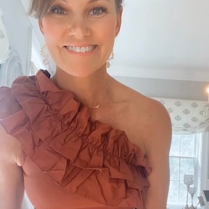 May include: A rust-colored one-shoulder top featuring a prominent, layered ruffle detail that extends across the chest and over one shoulder. The garment is constructed from a solid-colored, textured fabric with a fitted silhouette.