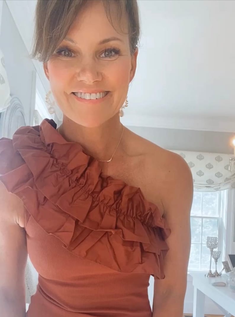 May include: A rust-colored one-shoulder top featuring a prominent, layered ruffle detail that extends across the chest and over one shoulder. The garment is constructed from a solid-colored, textured fabric with a fitted silhouette.