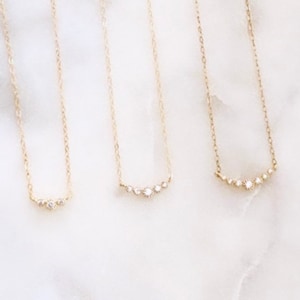 May include: Three gold necklaces with delicate chains, each featuring a small horizontal bar pendant set with tiny clear stones. The first necklace has three stones, the second has four, and the third has five stones arranged in a curved line.