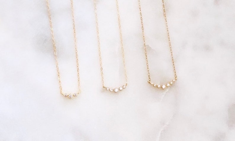 May include: Three gold necklaces with delicate chains, each featuring a small horizontal bar pendant set with tiny clear stones. The first necklace has three stones, the second has four, and the third has five stones arranged in a curved line.