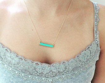 May include: A gold-toned necklace featuring a horizontal rectangular bar pendant with a bright turquoise inlay. The pendant is suspended from a delicate gold-toned chain that rests against the collarbone area of the wearer.