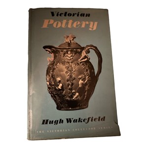 May include: A blue book cover with the title "Victorian Pottery" in black text. The cover features a detailed image of a brown and gold ceramic pitcher with ornate designs and figures. The author's name, "Hugh Wakefield", is printed below the image, along with the text "The Victorian Collector Series".
