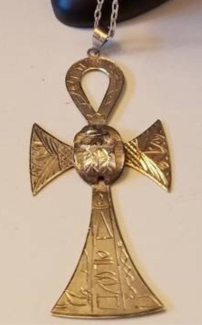 May include: A gold-toned Ankh pendant necklace with intricate details and a cross design. The pendant features a raised, circular design in the center.