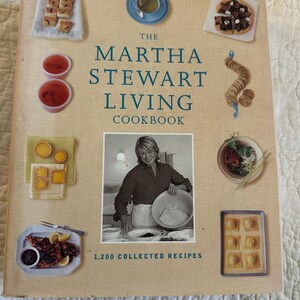 May include: The Martha Stewart Living Cookbook, 1,200 Collected Recipes, features a beige cover with a central black and white photograph of a person mixing ingredients in a bowl, surrounded by various color images of prepared food dishes.
