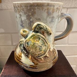 May include: A ceramic mug with a sculpted turtle design. The mug features a light blue and brown glaze with a textured floral pattern. The turtle is detailed with green, yellow, and brown hues.