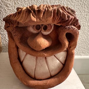 May include: A whimsical, terracotta-colored ceramic face with a wide, toothy grin. The sculpture features exaggerated facial features, including large eyes, a prominent nose, and textured hair. The piece has a rustic, handmade appearance.