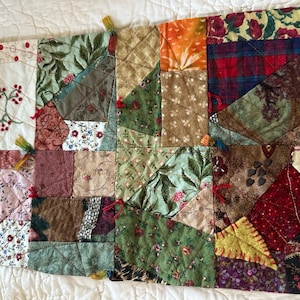May include: A rectangular patchwork quilt featuring a variety of fabric squares in shades of green, red, brown, and yellow. The piece includes floral, plaid, and solid patterns with decorative stitching, embroidery, and small tassels at the corners.
