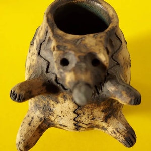 May include: A small, brown ceramic planter shaped like a bear with a textured surface. The planter has a round opening at the top and four legs.